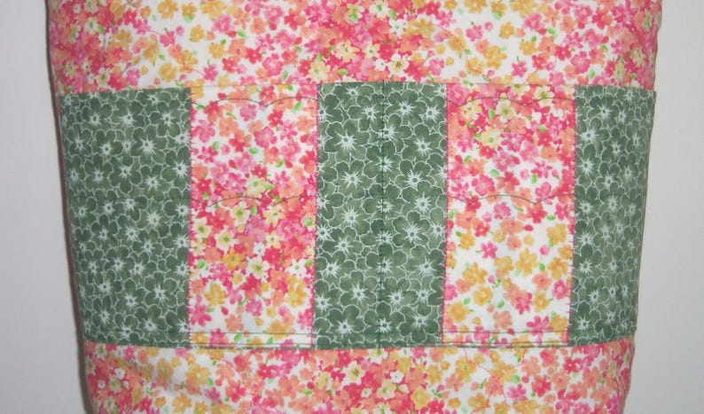 May include: A fabric bag featuring a repeating floral pattern in shades of pink, orange, and yellow on a white background. The exterior includes a horizontal band of alternating vertical panels of green floral fabric and pink floral fabric.
