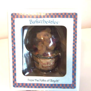 Op de afbeelding: Een beeldje in een doos van "Basketbearies" met een beer die een fruitmand vasthoudt. De beer is lichtbruin en de mand heeft de tekst "Thanks a Bushel". De doos heeft een blauw-oranje geruite rand.