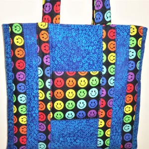 Puede incluir: Un bolso tote confeccionado con paneles de tela acolchada que presenta un patr&oacute;n repetitivo de caras sonrientes multicolores sobre un fondo negro, alternado con tiras verticales de tela azul con un estampado animal texturizado.