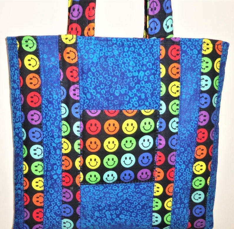 Puede incluir: Un bolso tote confeccionado con paneles de tela acolchada que presenta un patr&oacute;n repetitivo de caras sonrientes multicolores sobre un fondo negro, alternado con tiras verticales de tela azul con un estampado animal texturizado.
