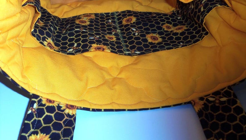 May include: A quilted fabric bag interior featuring a bright yellow lining and a black pocket panel decorated with a repeating pattern of yellow sunflowers and honeycombs. The bag has a soft, padded texture with visible stitching throughout.