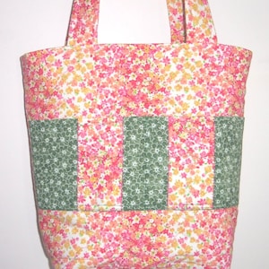 May include: A quilted tote bag featuring a vibrant floral pattern in shades of pink, orange, and yellow. The exterior includes three vertical panels of dark green fabric with a white floral print, creating distinct pockets across the front.