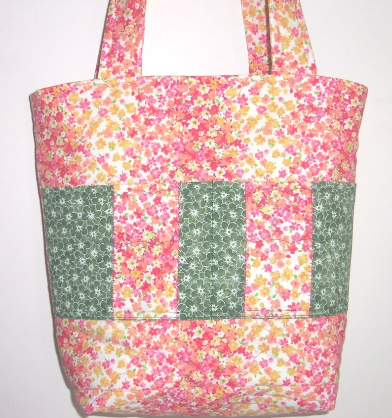 May include: A quilted tote bag featuring a vibrant floral pattern in shades of pink, orange, and yellow. The exterior includes three vertical panels of dark green fabric with a white floral print, creating distinct pockets across the front.
