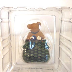 May include: A small, brown teddy bear figurine wearing a blue collar, seated inside a green and black woven basket. The bear is packaged in a clear plastic container, ready for display or gifting.