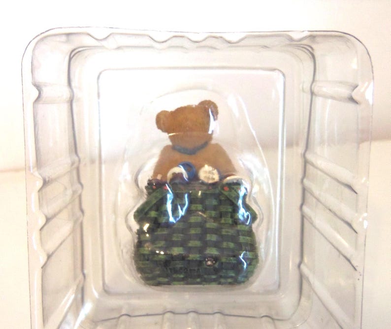 May include: A small, brown teddy bear figurine wearing a blue collar, seated inside a green and black woven basket. The bear is packaged in a clear plastic container, ready for display or gifting.