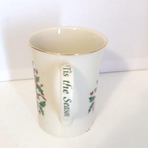 May include: A white ceramic mug featuring a gold-trimmed rim and a handle printed with the green text 'Tis the Season'. The mug is decorated with a festive holly and berry pattern, showcasing green leaves and red berries on its exterior surface.