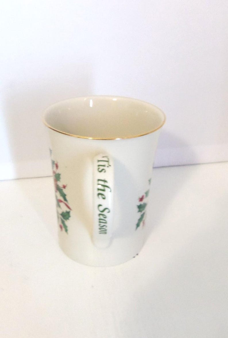 May include: A white ceramic mug featuring a gold-trimmed rim and a handle printed with the green text 'Tis the Season'. The mug is decorated with a festive holly and berry pattern, showcasing green leaves and red berries on its exterior surface.