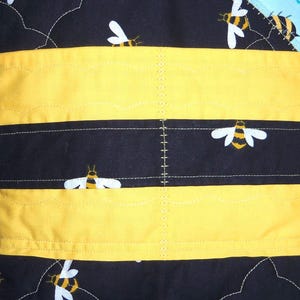 K&ouml;nnte beinhalten: Eine gesteppte Stofftasche mit schwarzem Hintergrund und einem sich wiederholenden Muster aus gelben und schwarzen Bienen. Das Design umfasst horizontale gelbe Stoffb&auml;nder und dekorative wei&szlig;e N&auml;hte auf der gesamten gesteppten Oberfl&auml;che.