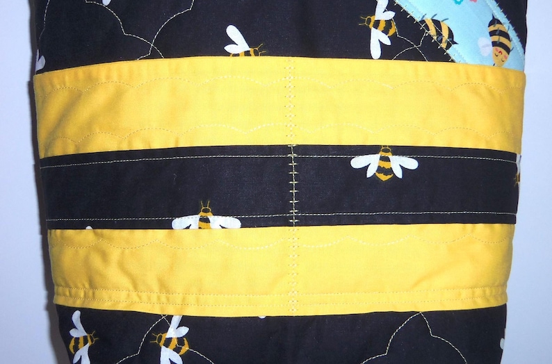 K&ouml;nnte beinhalten: Eine gesteppte Stofftasche mit schwarzem Hintergrund und einem sich wiederholenden Muster aus gelben und schwarzen Bienen. Das Design umfasst horizontale gelbe Stoffb&auml;nder und dekorative wei&szlig;e N&auml;hte auf der gesamten gesteppten Oberfl&auml;che.