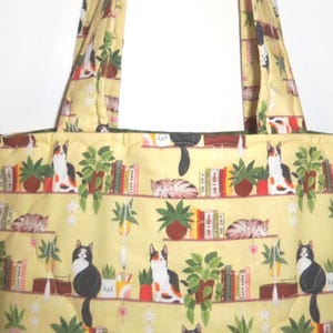 May include: A quilted tote bag in a pale yellow fabric featuring a repeating pattern of cats, potted green plants, and books arranged on shelves. The bag includes two matching fabric shoulder straps and a stitched grid texture throughout the surface.