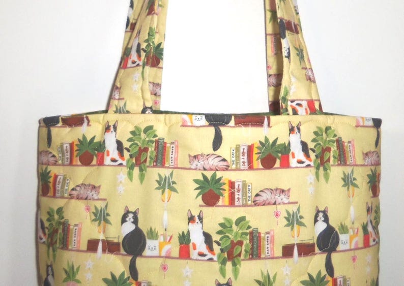 May include: A quilted tote bag in a pale yellow fabric featuring a repeating pattern of cats, potted green plants, and books arranged on shelves. The bag includes two matching fabric shoulder straps and a stitched grid texture throughout the surface.