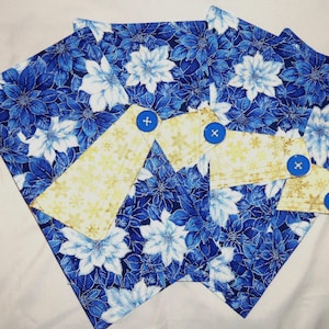 May include: A set of fabric items featuring a blue and white floral pattern with poinsettia-like flowers. Overlaid are three light yellow fabric tabs with a gold snowflake print, each secured with a single bright blue plastic button.