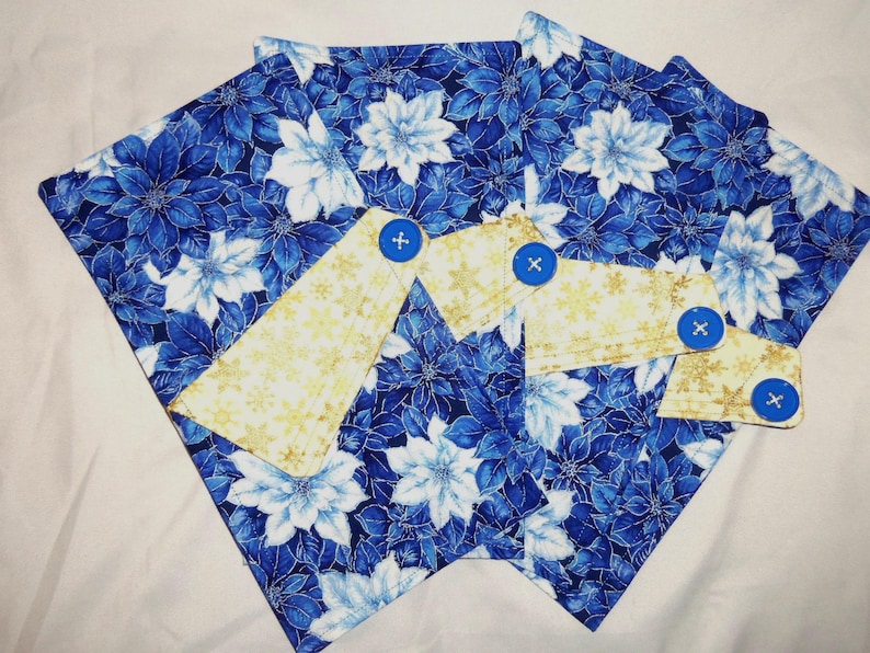 May include: A set of fabric items featuring a blue and white floral pattern with poinsettia-like flowers. Overlaid are three light yellow fabric tabs with a gold snowflake print, each secured with a single bright blue plastic button.