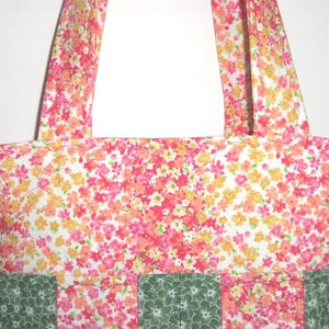 May include: A tote bag featuring a floral pattern with pink, orange, and yellow blossoms on a white background. The lower section of the bag is constructed with alternating vertical panels of the floral fabric and a dark green fabric with a white floral print.