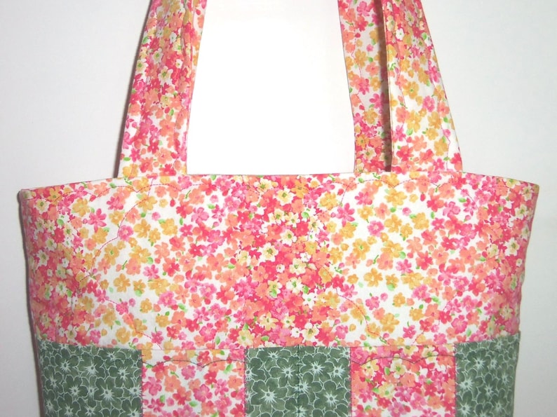 May include: A tote bag featuring a floral pattern with pink, orange, and yellow blossoms on a white background. The lower section of the bag is constructed with alternating vertical panels of the floral fabric and a dark green fabric with a white floral print.