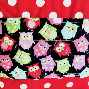 May include: A quilted fabric item featuring a red background with white polka dots and a central horizontal black band decorated with a repeating pattern of colourful owls in shades of pink, green, and blue, with the word snooz printed on some.