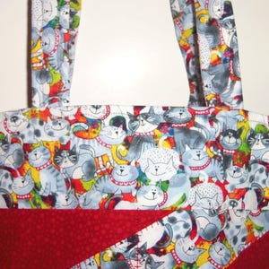 May include: A fabric tote bag featuring a repeating pattern of cartoon cats in shades of gray, white, and black against a colorful background. The bag includes two matching fabric handles and a solid dark red fabric panel at the bottom section.