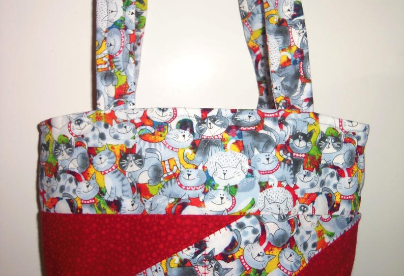 May include: A fabric tote bag featuring a repeating pattern of cartoon cats in shades of gray, white, and black against a colorful background. The bag includes two matching fabric handles and a solid dark red fabric panel at the bottom section.