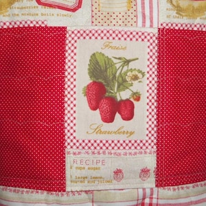 May include: A quilted fabric panel featuring a central rectangular patch with a botanical illustration of strawberries, the French word Fraise, and the English word Strawberry. The patch includes recipe text and red polka dot side panels.