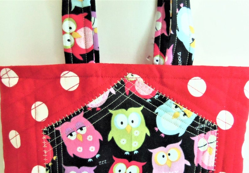 May include: A quilted tote bag featuring a vibrant red fabric with white polka dots and a black front pocket decorated with a colourful owl pattern. The bag includes matching black fabric handles with an owl print and white stitched details.