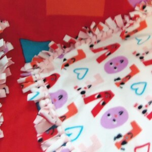 May include: A red and white fleece blanket with a knotted fringe. The blanket features the word "LOVE" in red, along with purple faces and blue hearts. The blanket is folded over a gray surface.