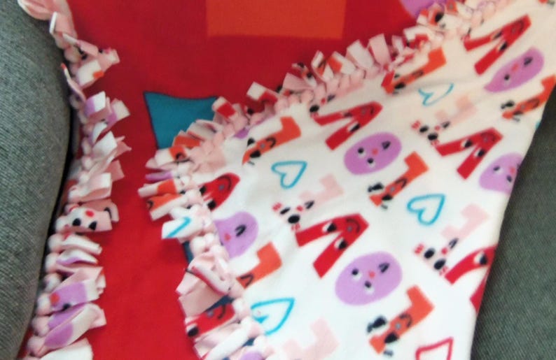 May include: A red and white fleece blanket with a knotted fringe. The blanket features the word "LOVE" in red, along with purple faces and blue hearts. The blanket is folded over a gray surface.