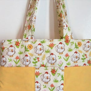 May include: A quilted tote bag featuring a repeating pattern of white cartoon chickens, green grass, and orange flowers on a cream background, with solid yellow fabric panels at the bottom and two matching patterned fabric shoulder straps.