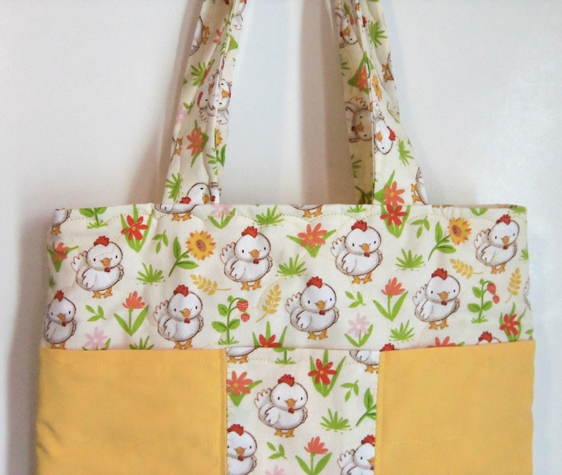 May include: A quilted tote bag featuring a repeating pattern of white cartoon chickens, green grass, and orange flowers on a cream background, with solid yellow fabric panels at the bottom and two matching patterned fabric shoulder straps.