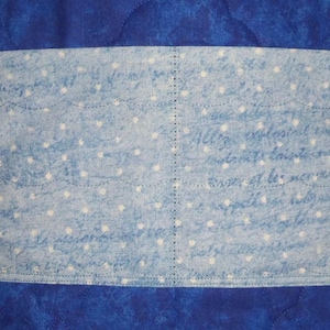 May include: A quilted fabric item featuring a deep blue marbled background and a central horizontal band of light blue fabric. The band is decorated with a white polka dot pattern and faint, illegible cursive script, finished with visible stitching.