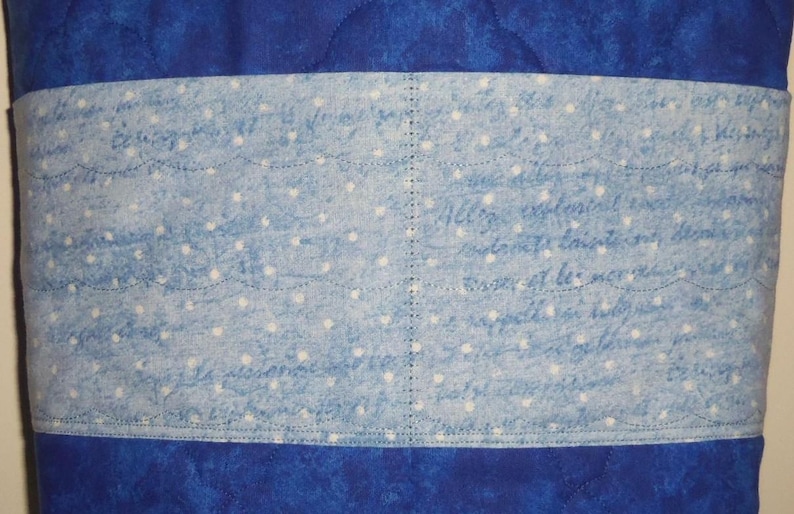May include: A quilted fabric item featuring a deep blue marbled background and a central horizontal band of light blue fabric. The band is decorated with a white polka dot pattern and faint, illegible cursive script, finished with visible stitching.