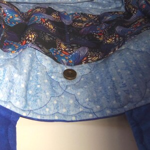 May include: A quilted fabric tote bag interior featuring a light blue lining with white polka dots and a scalloped stitching pattern. A dark fabric band with a colorful butterfly print runs along the top edge, secured by a central magnetic snap.