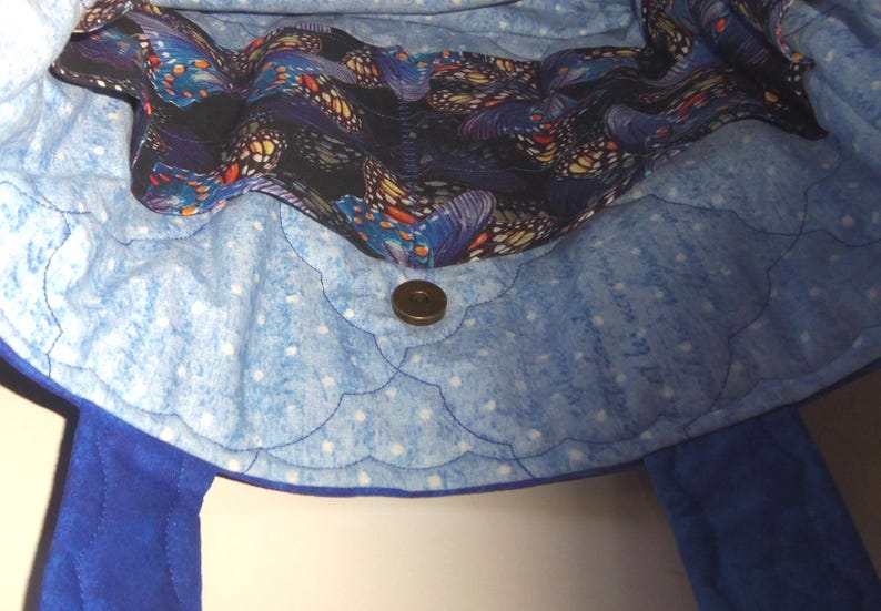 May include: A quilted fabric tote bag interior featuring a light blue lining with white polka dots and a scalloped stitching pattern. A dark fabric band with a colorful butterfly print runs along the top edge, secured by a central magnetic snap.
