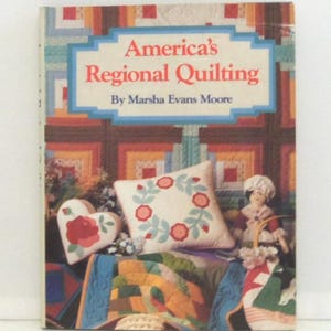 May include: A hardcover book titled America's Regional Quilting by Marsha Evans Moore. The cover features a colorful patchwork quilt background, a heart-shaped pillow with a red rose, a square pillow with a floral wreath, and a small fabric doll.