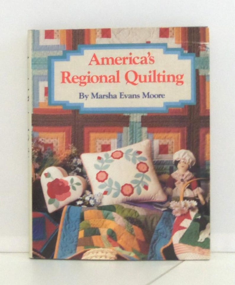 May include: A hardcover book titled America's Regional Quilting by Marsha Evans Moore. The cover features a colorful patchwork quilt background, a heart-shaped pillow with a red rose, a square pillow with a floral wreath, and a small fabric doll.