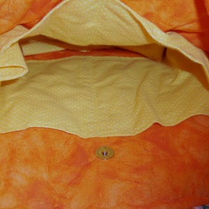 May include: Open view of a quilted tote bag interior. The bag features a vibrant orange exterior with a yellow polka-dot lining. A button closure is visible at the bottom. The bag's design suggests it's suitable for carrying items.