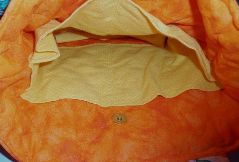 May include: Open view of a quilted tote bag interior. The bag features a vibrant orange exterior with a yellow polka-dot lining. A button closure is visible at the bottom. The bag's design suggests it's suitable for carrying items.