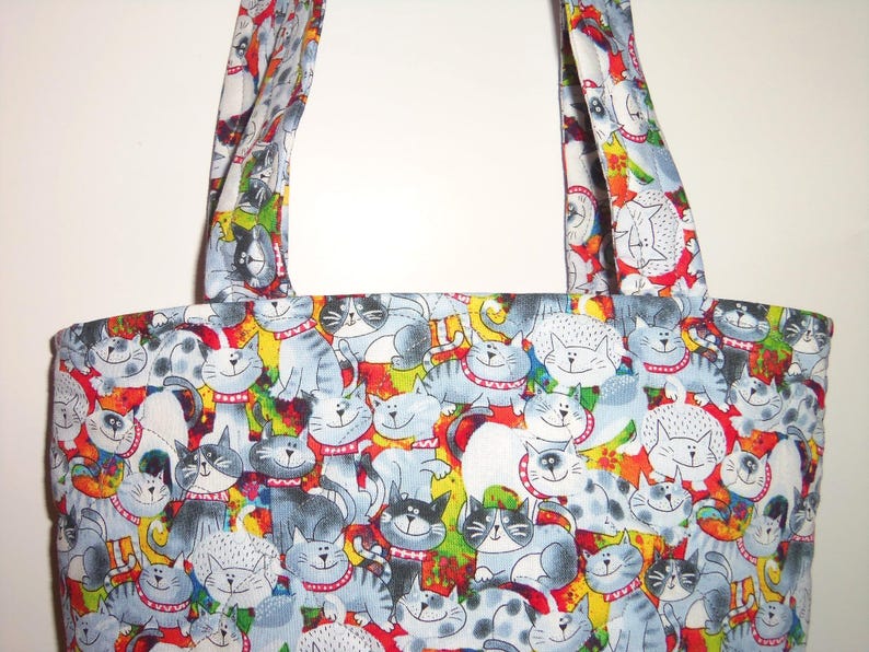 May include: A tote bag featuring a repeating pattern of cartoon cats in shades of gray, white, and black. The cats have expressive faces and wear red collars. The background includes vibrant splashes of yellow, green, and red colors throughout.