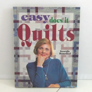 以下が含まれることがあります： ジョージア・ボーンスティール著のハードカバー本「Easy Does It Quilts」。表紙には、青いタートルネックのセーターと銀のネックレスを身につけた人物の写真が、白と青の格子模様のキルト柄の背景とともに印刷されています。