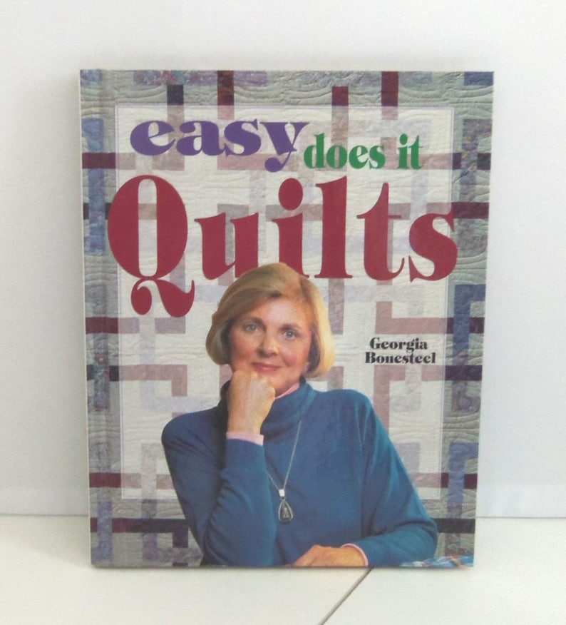 以下が含まれることがあります： ジョージア・ボーンスティール著のハードカバー本「Easy Does It Quilts」。表紙には、青いタートルネックのセーターと銀のネックレスを身につけた人物の写真が、白と青の格子模様のキルト柄の背景とともに印刷されています。