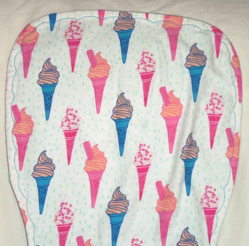 Op de afbeelding: Een witte baby slabbetje met geschulpte rand en een herhalend patroon van ijsjes. De ijsjes zijn roze en blauw, met bruin en oranje ijs, en sommige hebben hagelslag. Het slabbetje is afgezet met een blauwe steek.