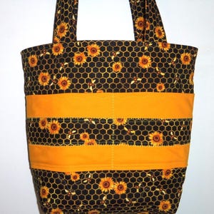 May include: A quilted tote bag featuring a black fabric with a yellow honeycomb pattern, sunflowers, and small bees. The bag includes two solid yellow horizontal bands across the center and two matching fabric shoulder straps at the top.