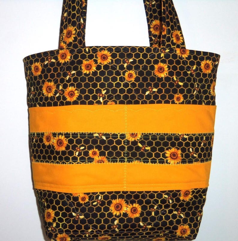 May include: A quilted tote bag featuring a black fabric with a yellow honeycomb pattern, sunflowers, and small bees. The bag includes two solid yellow horizontal bands across the center and two matching fabric shoulder straps at the top.