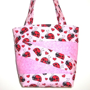 May include: A quilted tote bag featuring a light pink background with a repeating pattern of red ladybugs and small red hearts. The bag includes two shoulder straps and diagonal fabric panels with a subtle pink swirl texture integrated throughout.