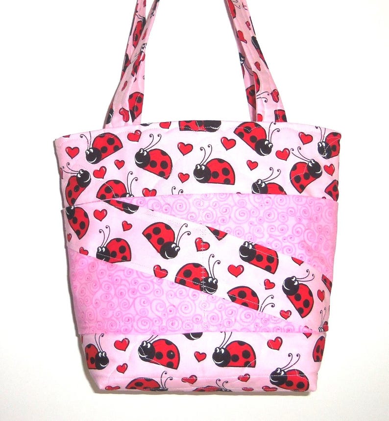 May include: A quilted tote bag featuring a light pink background with a repeating pattern of red ladybugs and small red hearts. The bag includes two shoulder straps and diagonal fabric panels with a subtle pink swirl texture integrated throughout.