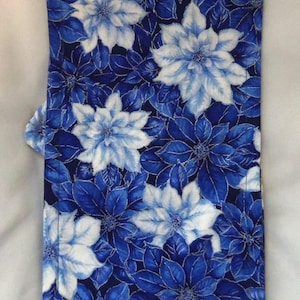 May include: A rectangular fabric piece featuring a repeating floral pattern of white and various shades of blue poinsettia-like flowers and leaves against a dark navy blue background, with visible stitching along the edges of the textile.
