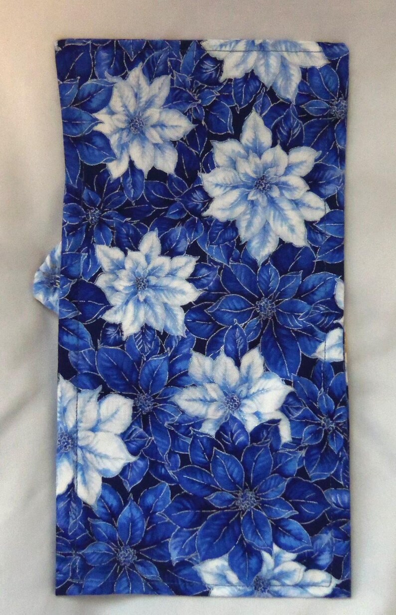 May include: A rectangular fabric piece featuring a repeating floral pattern of white and various shades of blue poinsettia-like flowers and leaves against a dark navy blue background, with visible stitching along the edges of the textile.