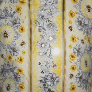May include: A fabric with a repeating pattern of bees, flowers, and crowns. The design features yellow, gray, and white elements, with a honeycomb-like background. The bees are detailed, and the flowers include daisies and other blooms.