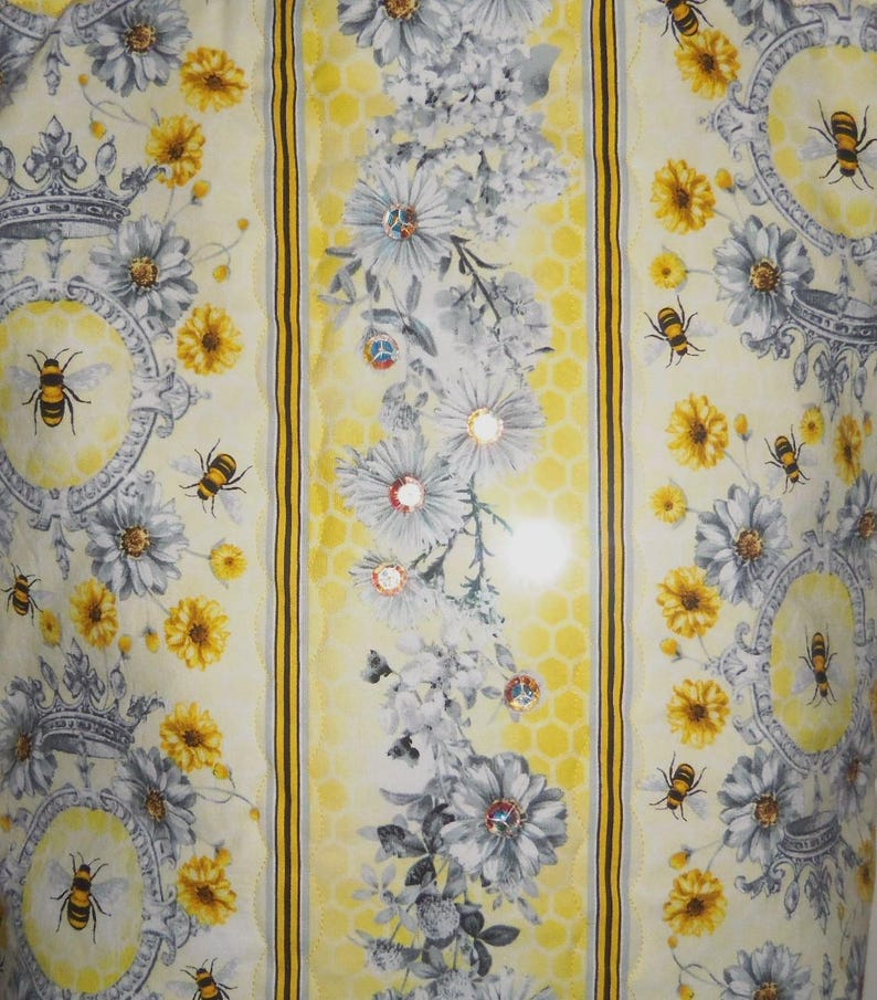 May include: A fabric with a repeating pattern of bees, flowers, and crowns. The design features yellow, gray, and white elements, with a honeycomb-like background. The bees are detailed, and the flowers include daisies and other blooms.