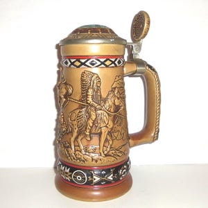 May include: A tan ceramic beer stein featuring a relief sculpture of a person in a feathered headdress riding a horse. The stein has a metal hinged lid, a sturdy handle, and decorative bands with geometric patterns in black, white, and red.