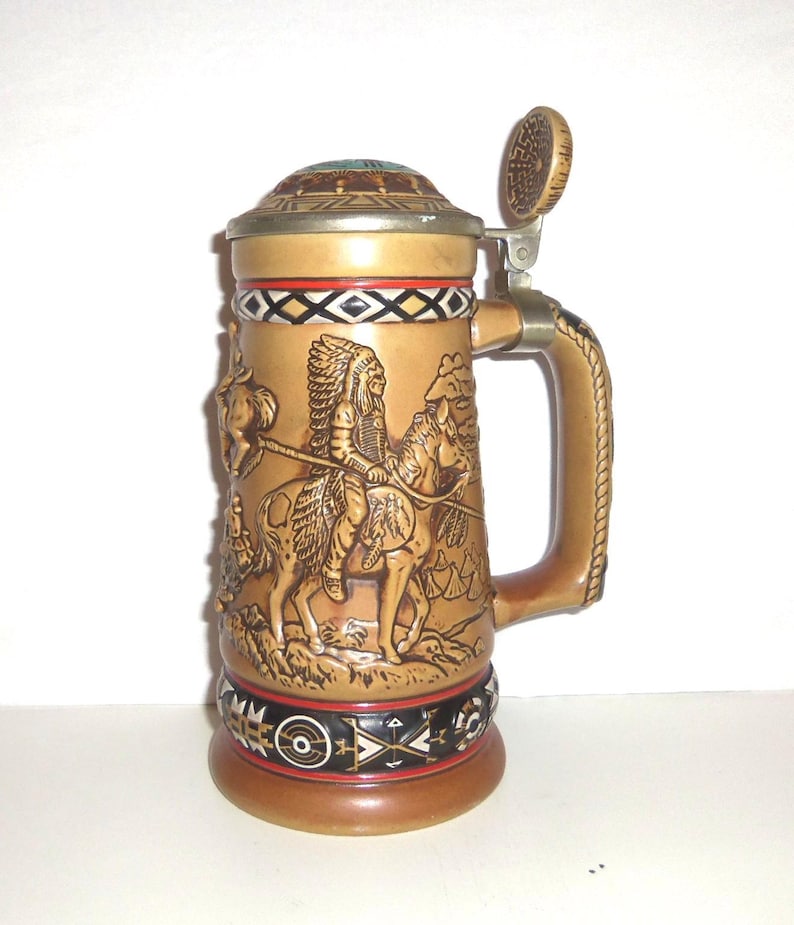May include: A tan ceramic beer stein featuring a relief sculpture of a person in a feathered headdress riding a horse. The stein has a metal hinged lid, a sturdy handle, and decorative bands with geometric patterns in black, white, and red.